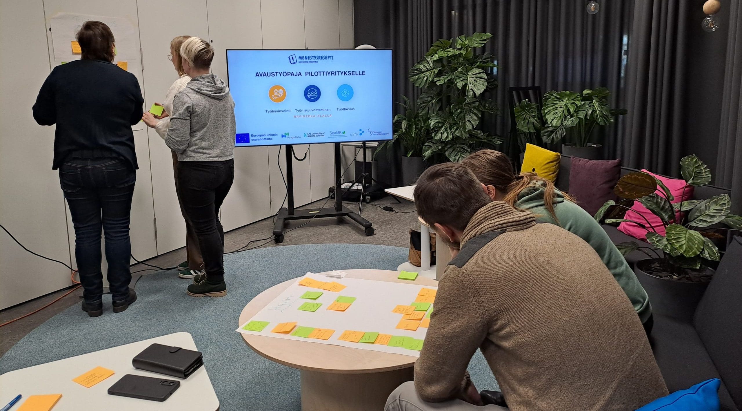 Henkilöitä ryhmissä keskittyneinä edessä oleviin papereihin ja post-it-lappuihin.