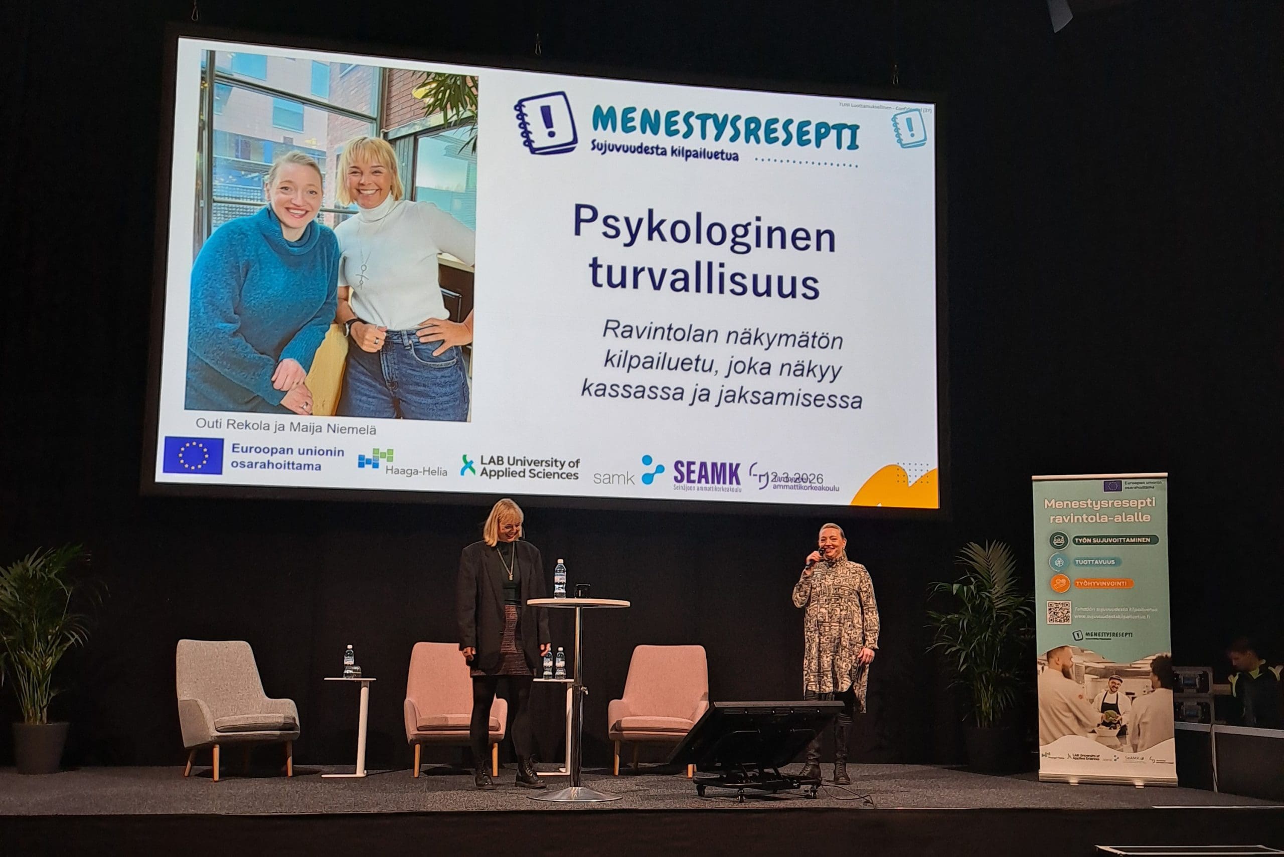 Maija ja Outi1 Kaksi naista lavalla seisomassa. Takan nojatuoleja sekä iso valkokangas, jossa lukee aihe: psykologinen turvallisuus.