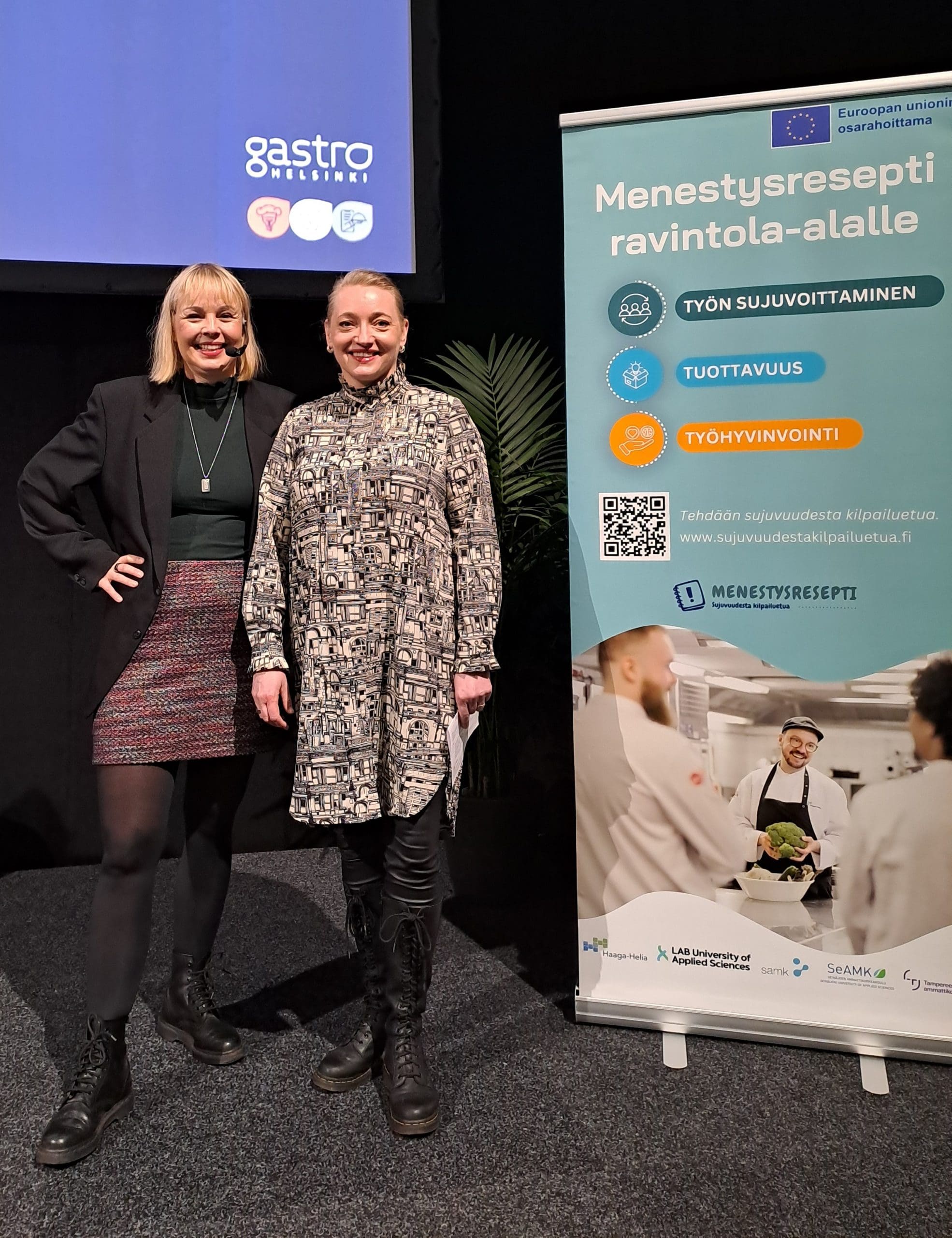 Kaksi naista hymyilemässä vierekkäin kameralle. Naisten vieressä on Menestysresepti-hankkeen roll-up.